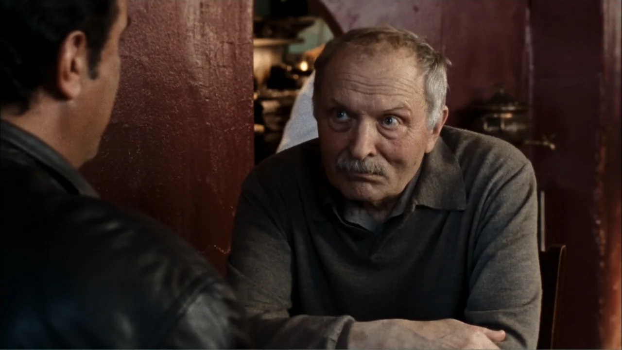 Driven to Kill (2009) movie still: medium — A dark restaurant booth features deep red walls and a wooden table. An older man with a mustache sits…