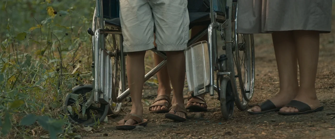 The Unholy (2022) movie still: medium — A dirt trail runs through a brushy area with vegetation on the left. Two individuals stand together while…