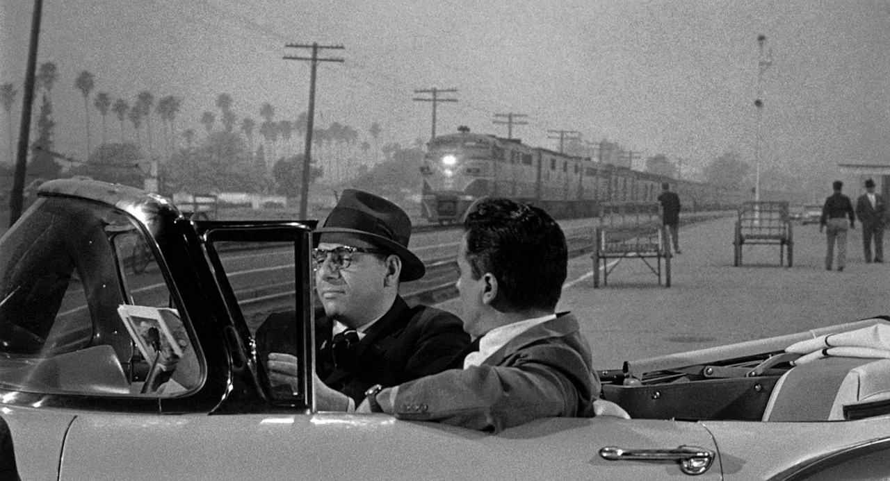 Murder By Contract (1958) movie still: medium — A flat railway station platform in the 1950s features train tracks and telephone poles. Two men sit…