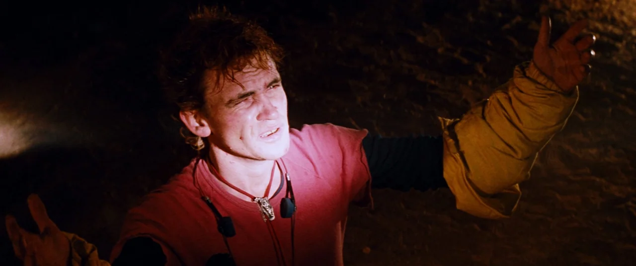 Razorback (1984) movie still: closeup — A dark, empty desert setting at night with no other structures visible. A young man gestures toward an…