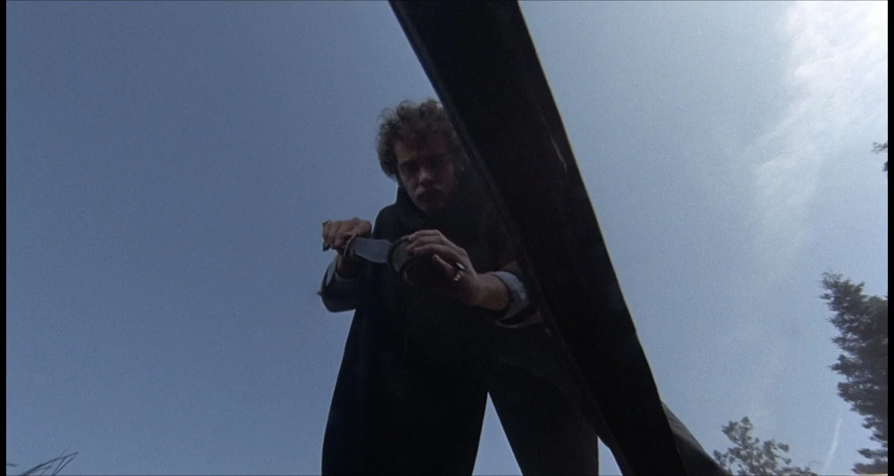 The Dunwich Horror (1970) movie still: medium — A figure stands in a bright outdoor field with clear blue sky visible. A young man with curly hair…