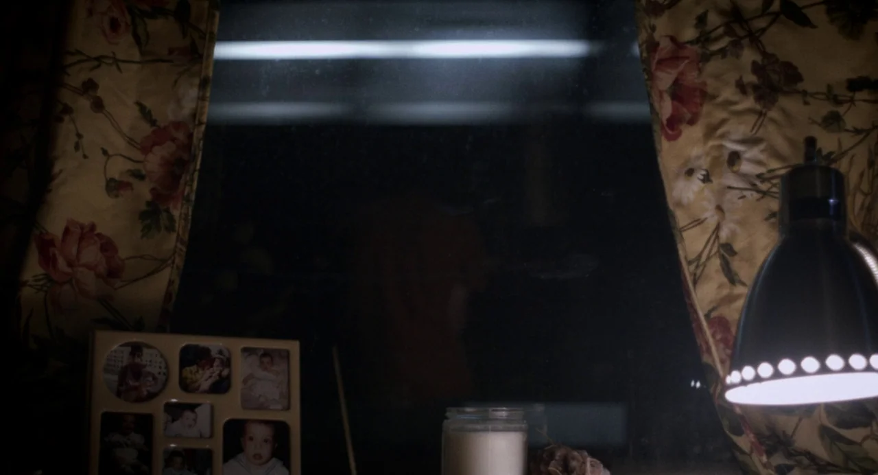 Happer’s Comet (2022) movie still: medium — A dark, dimly lit room features a window framed by floral patterned curtains. A photo collage frame sits…