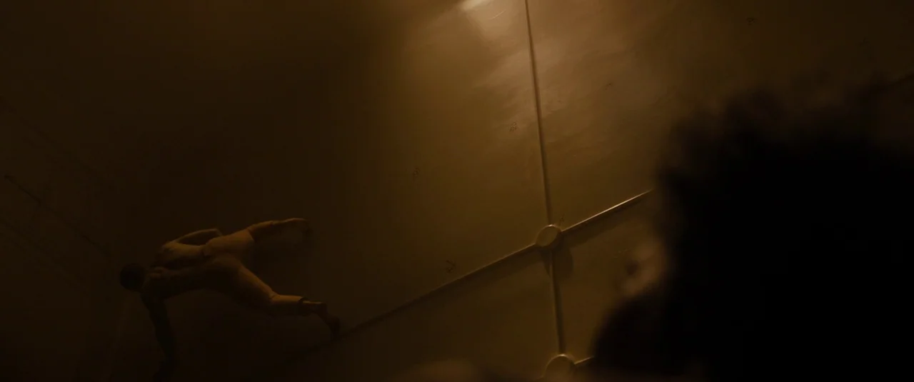 Glass (2019) movie still: wide — A dim interior corridor features smooth paneled walls with exposed structural junctions. A lone man crawls…