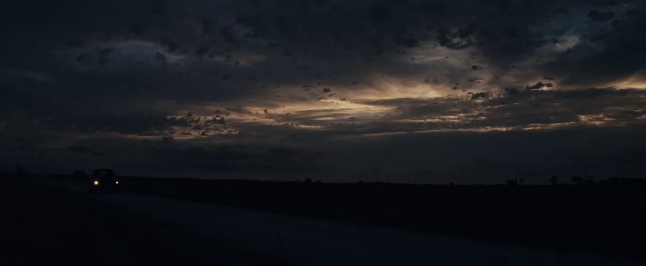 Ain’t Them Bodies Saints (2013) movie still: wide — A dark, expansive landscape stretches beneath a vast, cloudy sky at night. A lone vehicle drives…