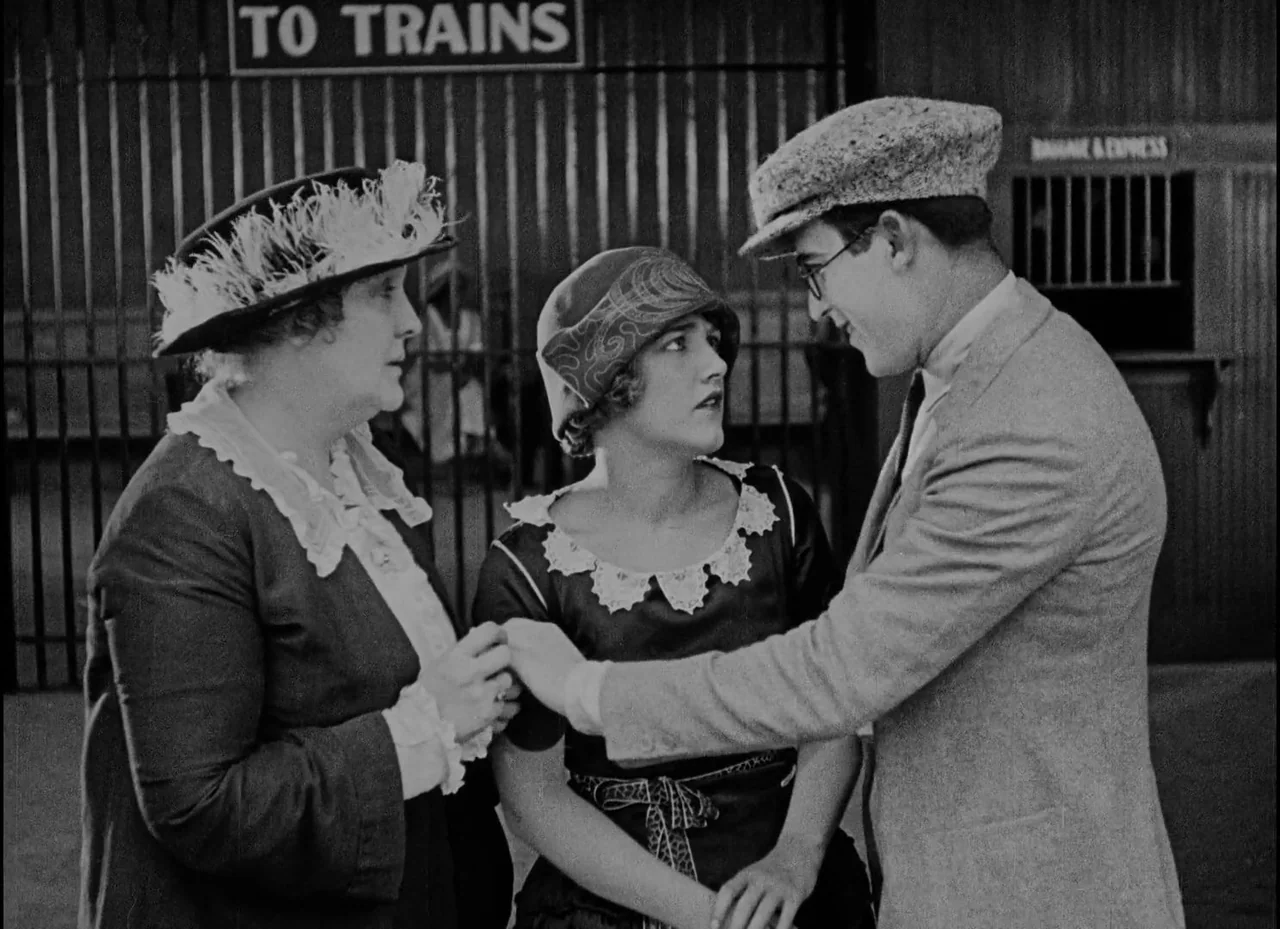 Safety Last! (1923) movie still: medium — A train station interior features a black gate with a To Trains sign. Three people stand together while…