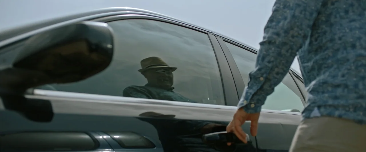 Hounds of War (2024) movie still: medium — A dark sedan sits in a bright outdoor space under a clear sky. A man reaches for the handle while the…