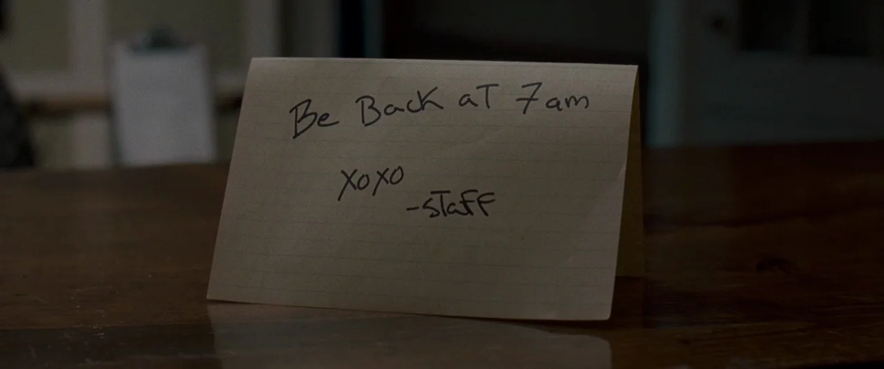 The Innkeepers (2011) movie still: closeup — A folded tan index card sits on a polished dark brown wooden surface. The card has handwritten text in…