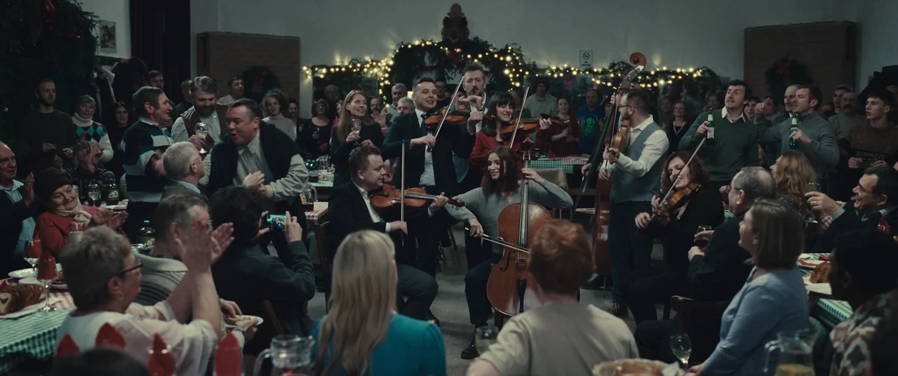 R.M.N. (2022) movie still: wide — A spacious community hall decorated with string lights hosts a gathering of many people. A string quartet stands…