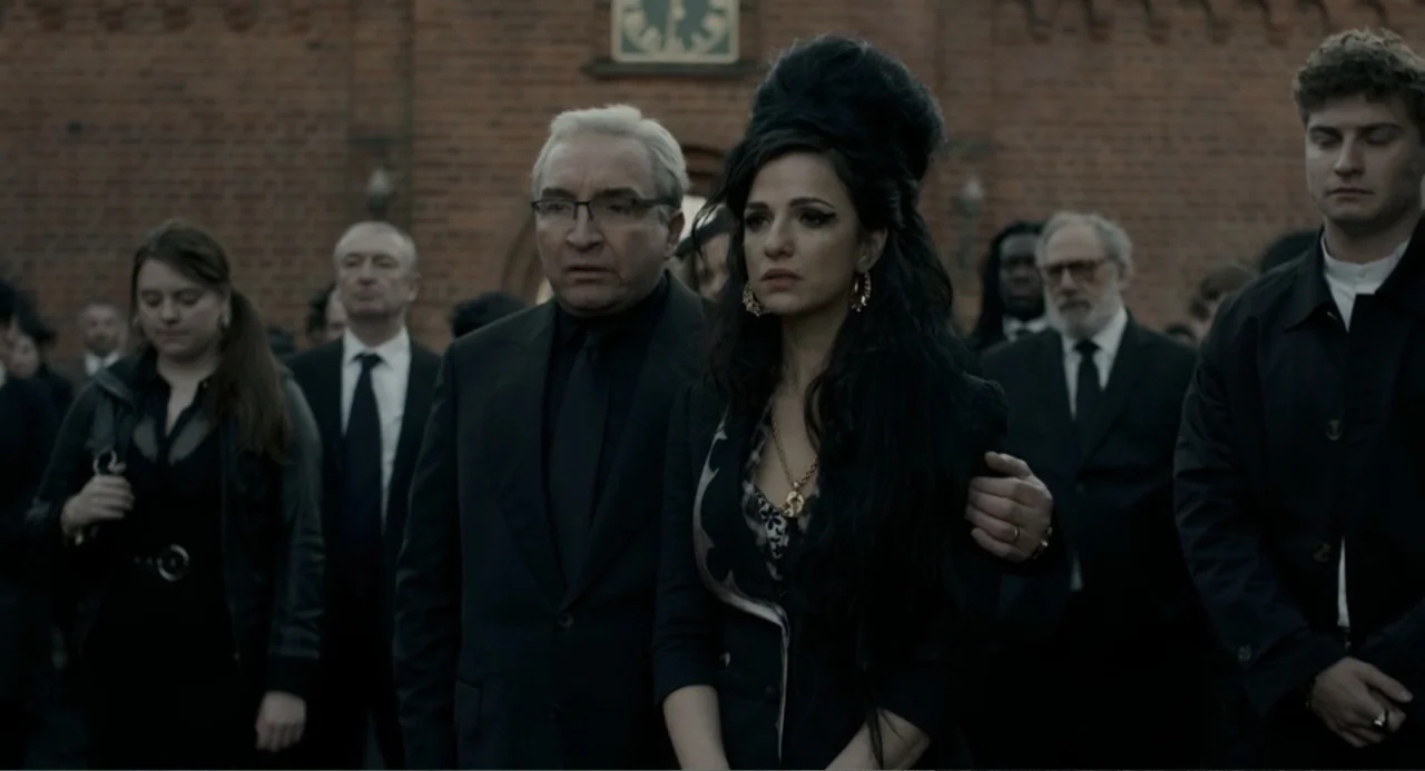 Back to Black (2024) movie still: medium — A group of mourners dressed in black stands before an old red brick building. A woman with a distinctive…