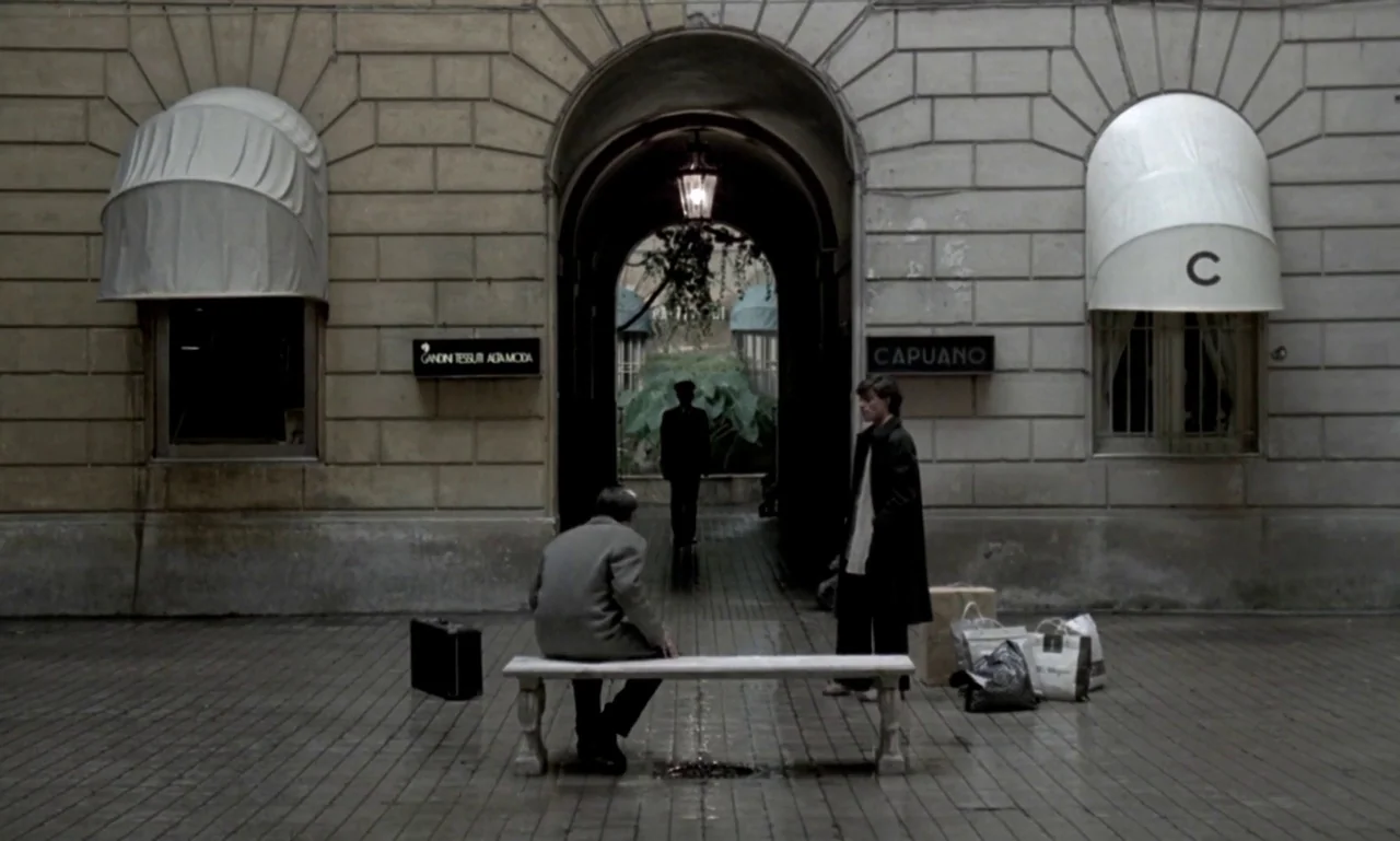 Nostalghia (1983) movie still: wide — A stone-paved building courtyard features two rounded fabric awnings and a central arched entryway. A man sits…