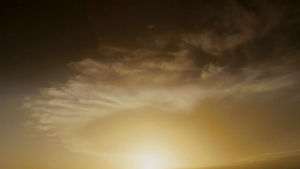 Sugar (2008) movie still: establishing — A vast sky is filled with swirling golden clouds at sunset. The sun glows brightly at the bottom center of…