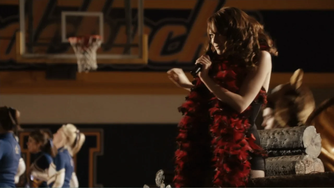 Easy A (2010) movie still: medium — A basketball court at night features a dark background with blurred sports bleachers. A young woman in a black…