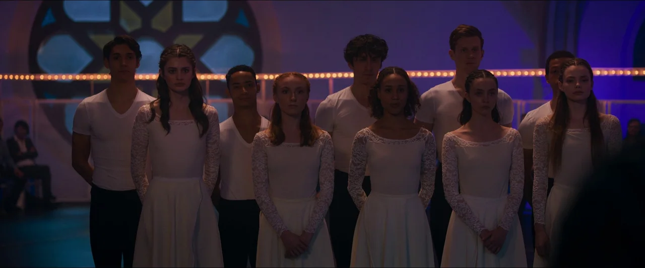 Birds of Paradise (2021) movie still: medium — A group of dancers in white uniform attire stands in formation inside a studio. A dark room features…