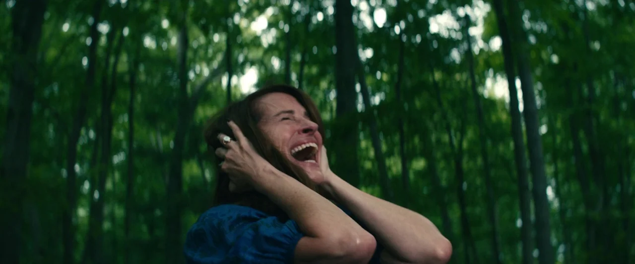 Leave The World Behind (2023) movie still: medium — A dense forest with tall trees blurred in the background beneath a canopy of leaves. A woman…