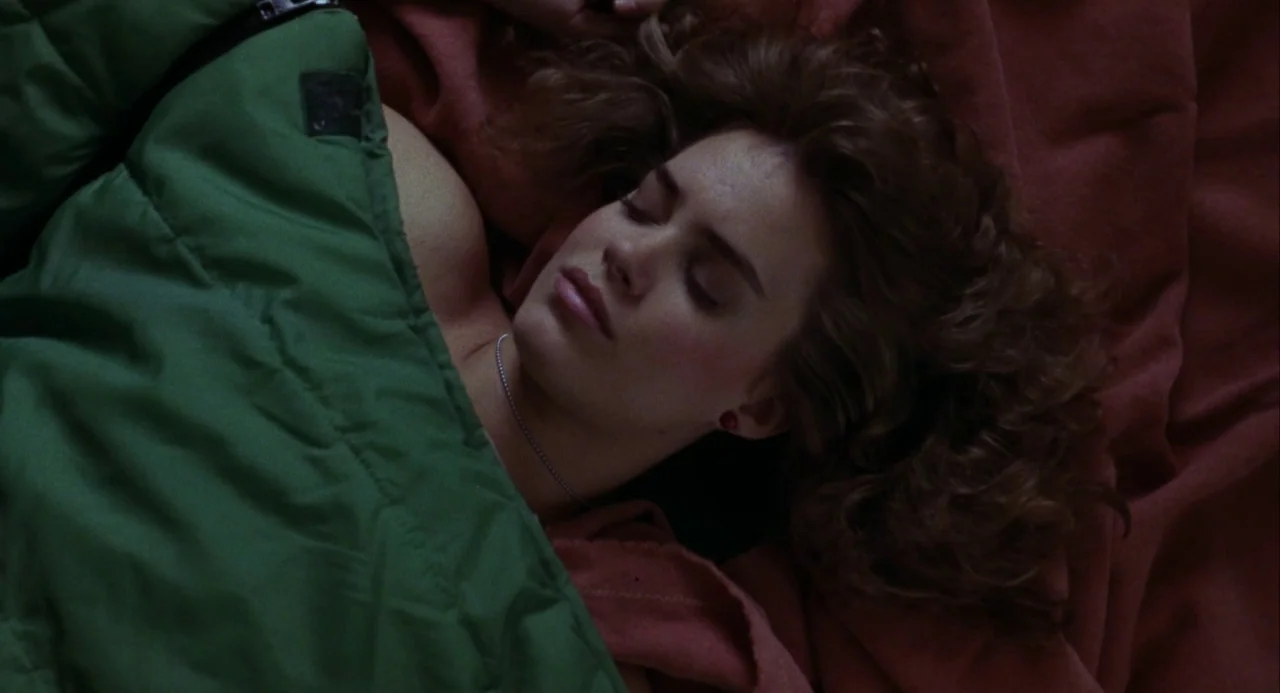 Night of the Comet movie still: closeup — A woman lies on a surface covered with reddish cloth and a green bag. She rests with eyes closed in a…