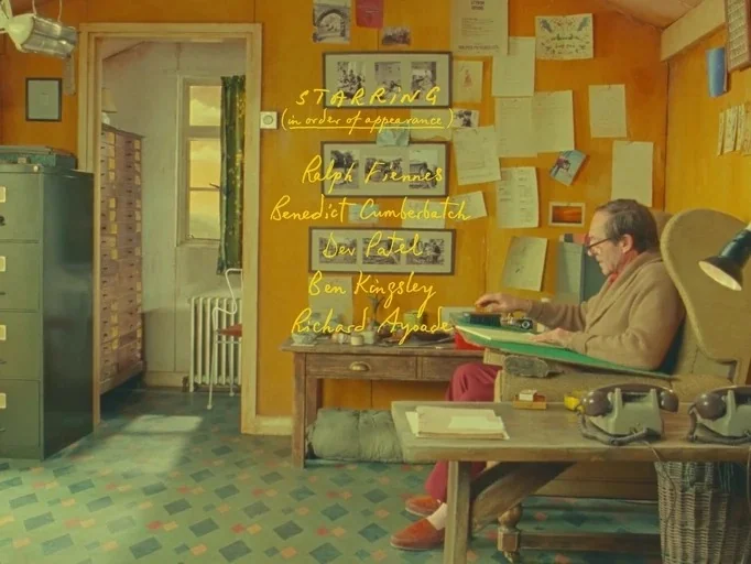 The Wonderful Story of Henry Sugar (2023) movie still: wide — A yellow-walled office with wooden furniture features numerous papers and framed…