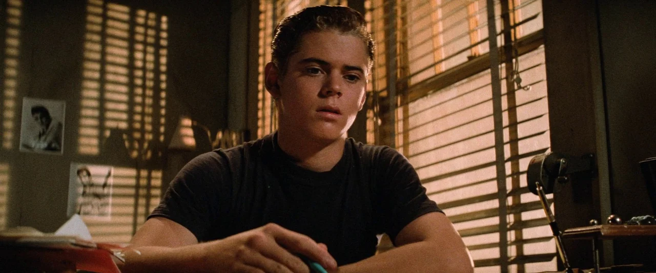 The Outsiders (1983) movie still: medium — A dark study features a desk positioned in front of a window with blinds. A teenage boy with slicked-back…