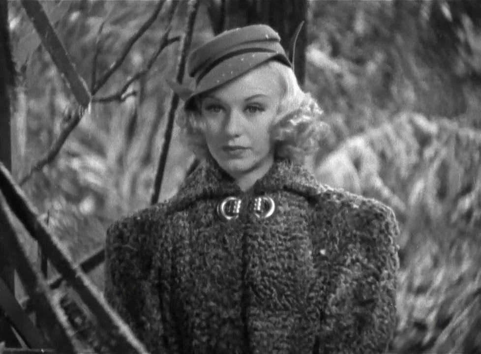 Swing Time (1936) movie still: medium — A woman stands among blurred trees and branches in a woodland setting. She wears a textured coat and a…
