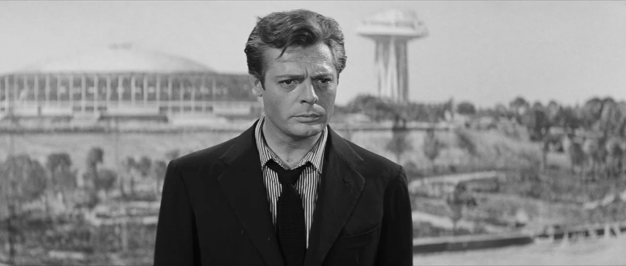 La Dolce Vita (1960) movie still: medium — A man stands in an outdoor park area with modern architectural structures visible in the distance. The…