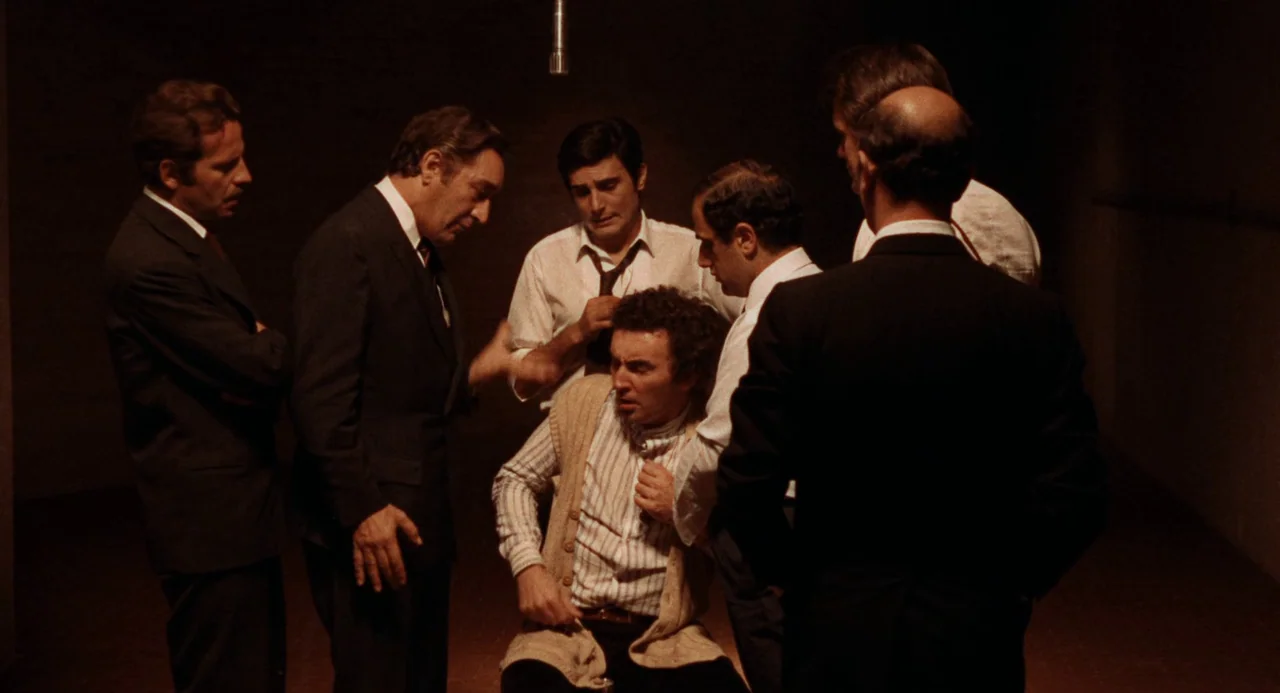 Investigation of a Citizen Above Suspicion (1970) movie still: medium — A dim basement room with concrete walls houses a group of men. Several men…