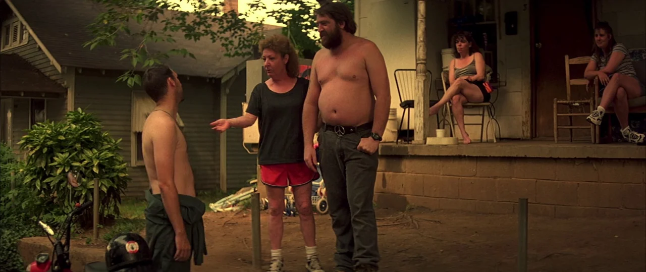 George Washington (2000) movie still: fullbody — A group of people gathers in the dirt yard in front of a house porch. The man stands shirtless…