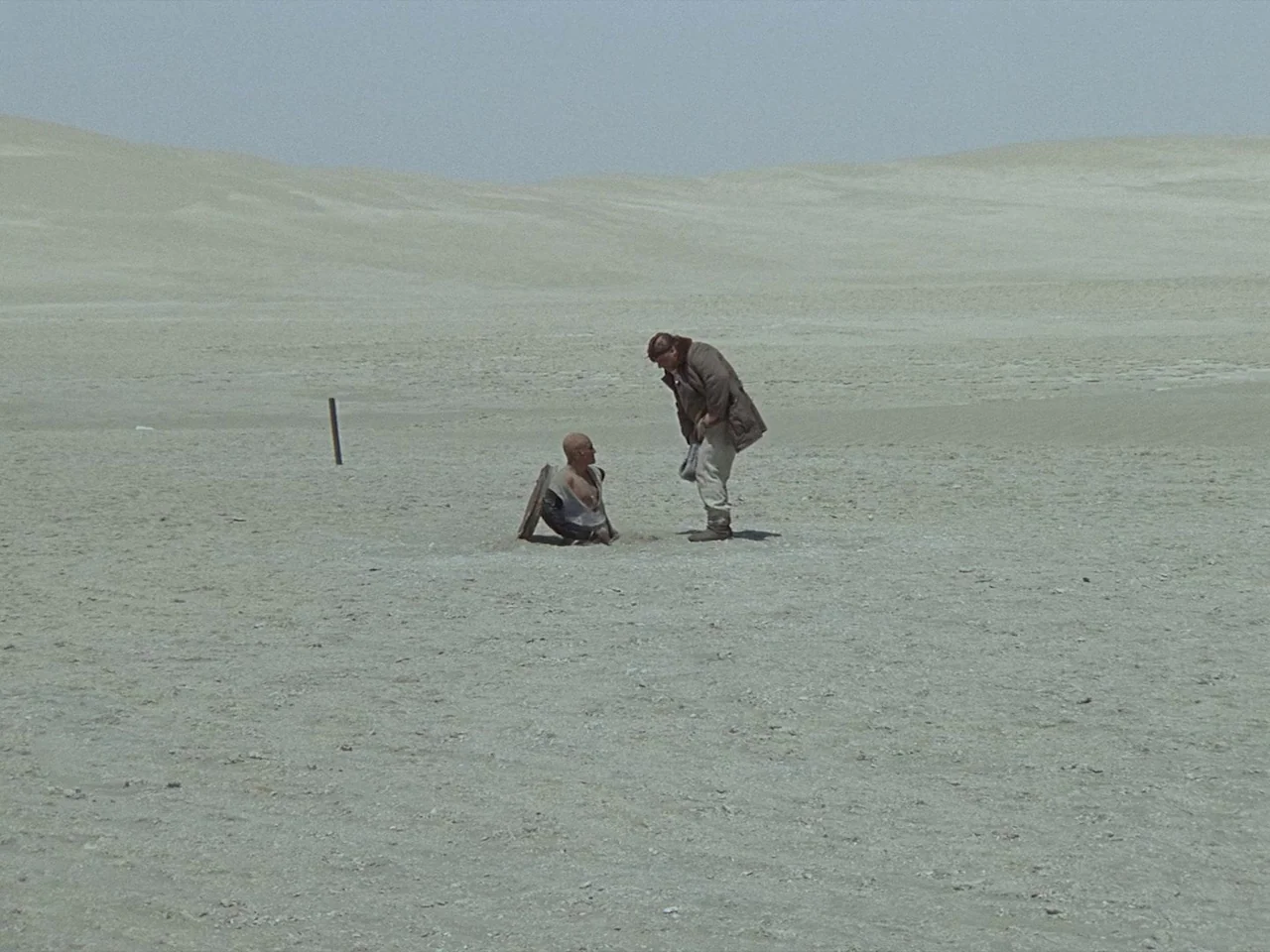 Kin-dza-dza! (1986) movie still: wide — A vast, barren desert landscape stretches to the horizon under a pale sky. A man stands over another man who…
