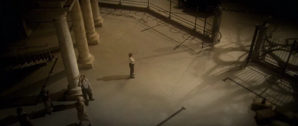 Manderlay (2005) movie still: wide — A stone courtyard features tall pillars and an ornate iron gate at night. A lone figure stands in the center…