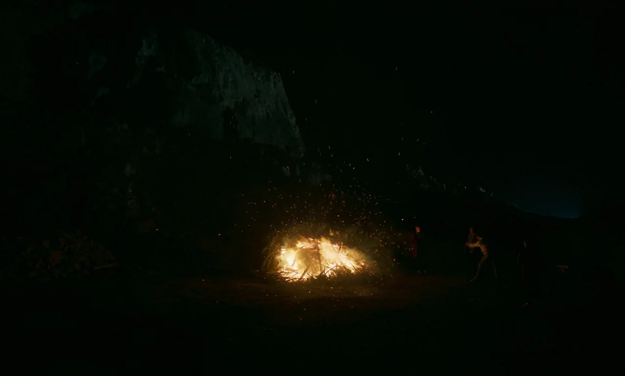 It Is In Us All (2022) movie still: wide — A dark, rocky quarry at night is illuminated by a crackling, large bonfire. Several figures stand…