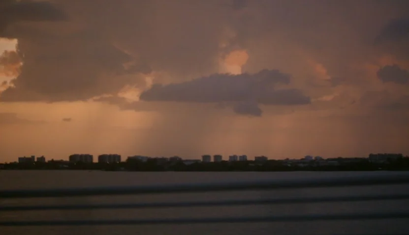 Kate Plays Christine (2016) movie still: establishing — A wide view of a calm body of water meeting a distant urban skyline. Low-hanging storm…