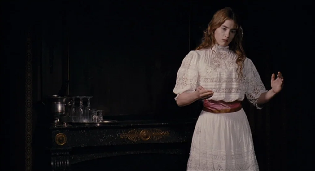 House of Tolerance (2011) movie still: medium — A dark, dimly lit room contains a marble fireplace topped with crystal glassware. A young woman in a…