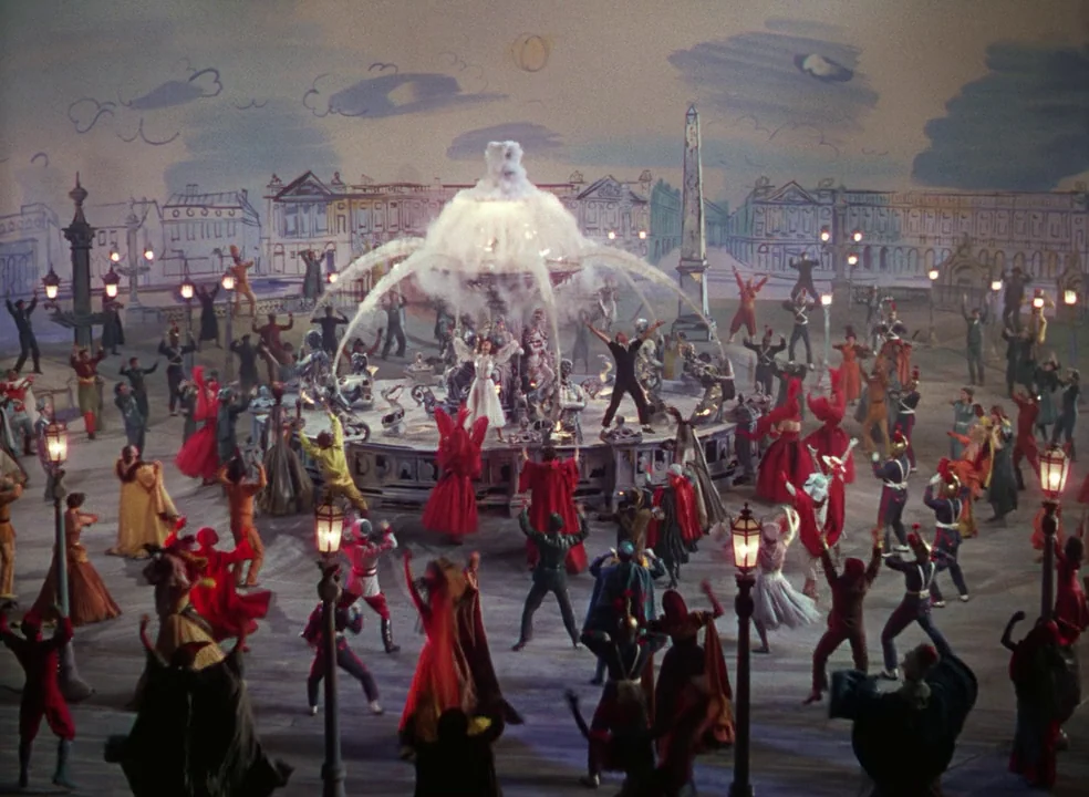 An American In Paris (1951) movie still: wide — A theatrical studio set features a painted backdrop of a Parisian square at night. A large crowd of…