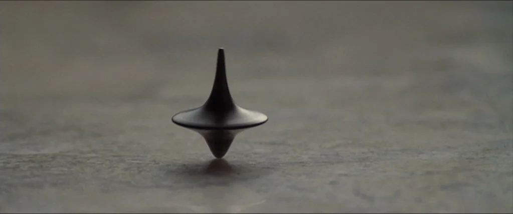 Inception (2010) movie still: closeup — A close-up shot focuses on a small metal top spinning on a smooth surface. The object is centered in the…