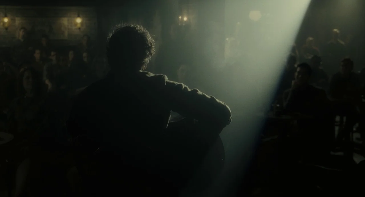 Inside Llewyn Davis (2013) movie still: medium — A dark, dimly lit venue with a seated audience watching a performer. A musician sits on a chair,…