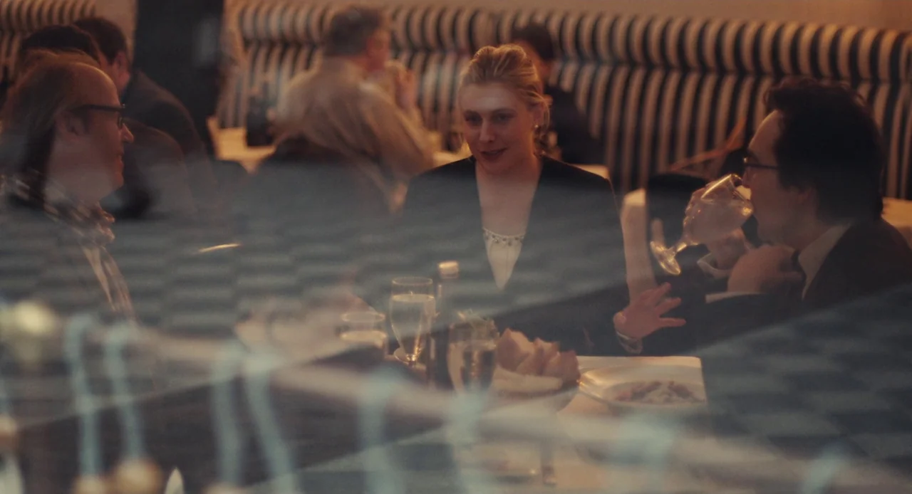 Mistress America (2015) movie still: medium — A group of three people converse around a dining table inside a restaurant. The scene is shot through…