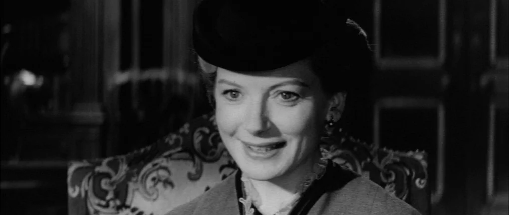 The Innocents (1961) movie still: closeup — A dimly lit room features a woman seated in a patterned upholstered chair. She wears a formal hat while…