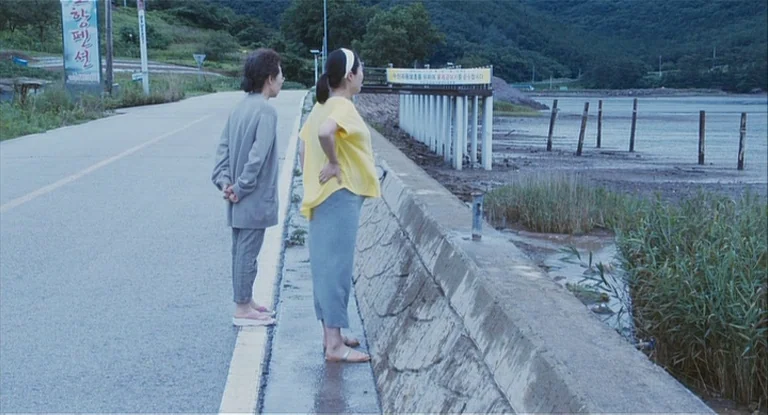 In Another Country (2012) movie still: fullbody — A paved coastal road bordered by a concrete sea wall and tall marsh grass. Two women stand…