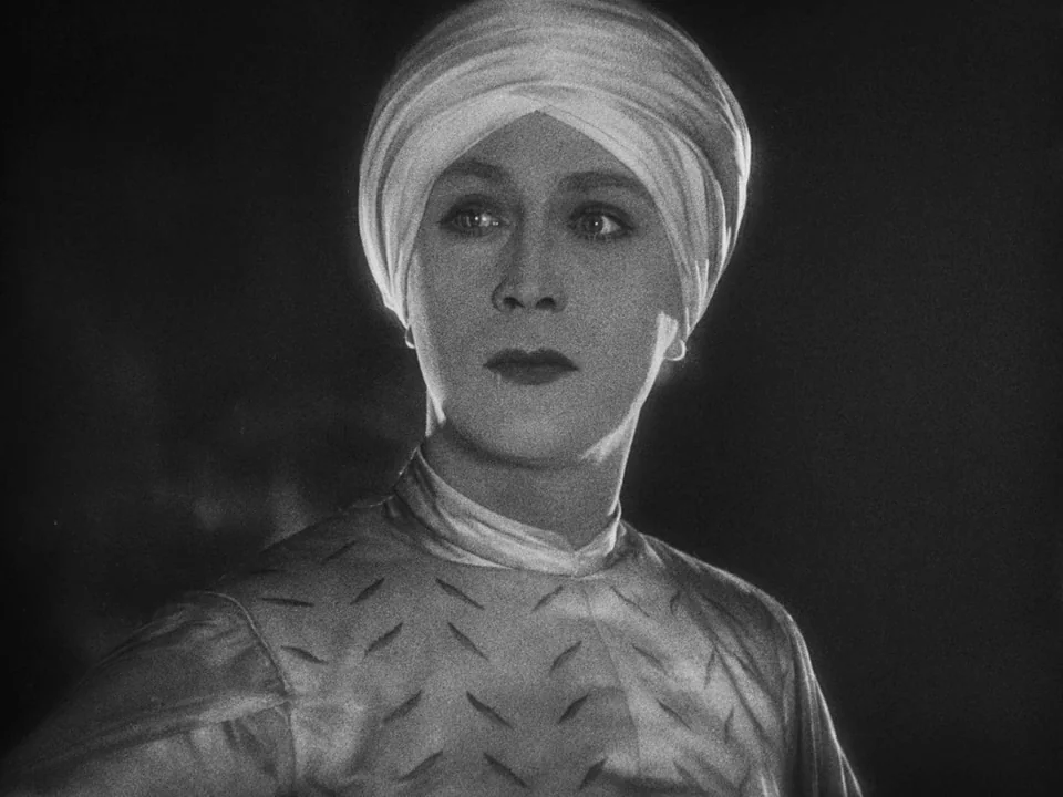 Faust (1926) movie still: closeup — A head and shoulders portrait of a woman wearing a white head wrap. The figure is positioned against a plain…