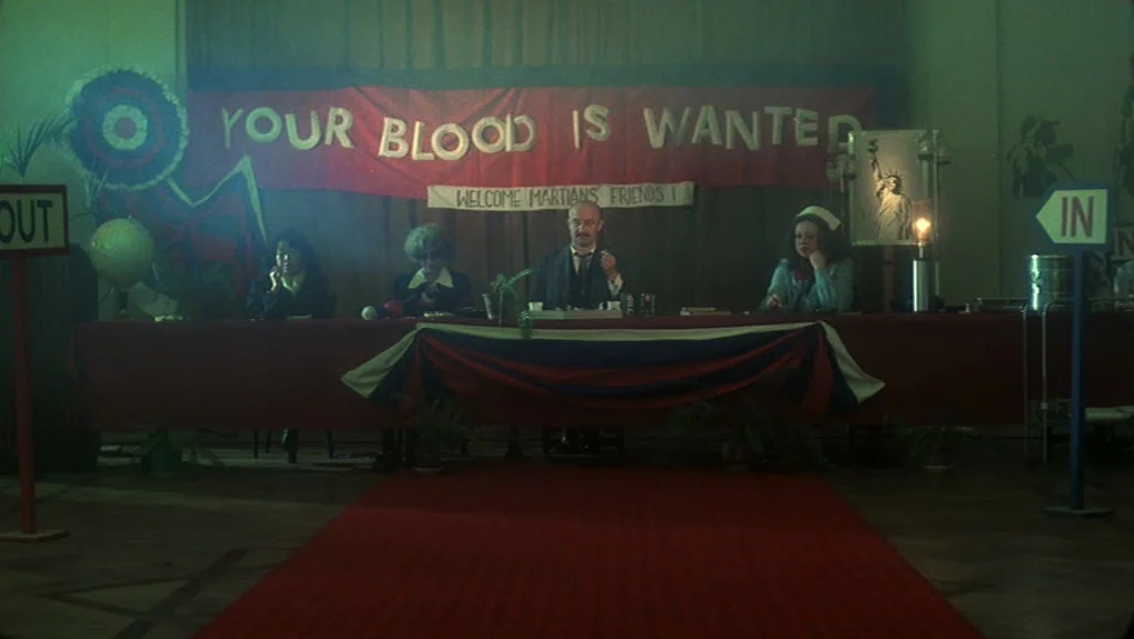 The War of the Worlds: Next Century (1981) movie still: wide — A dimly lit hall features a long table with four people seated behind it. A large red…