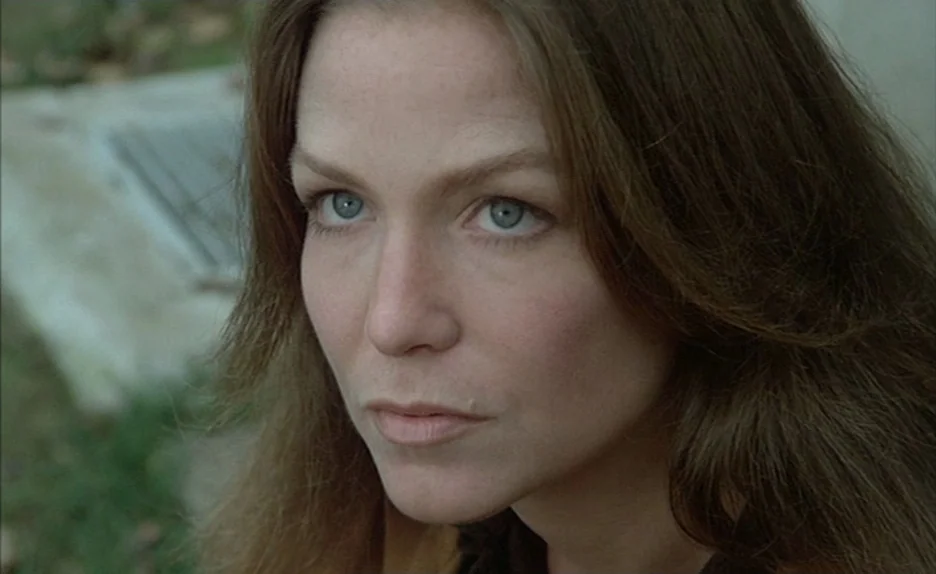 Black Moon (1975) movie still: closeup — A close-up view shows a woman's face against a blurred outdoor grass background. The subject gazes forward…