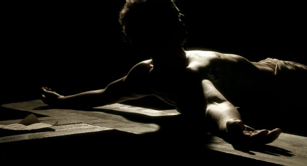 The Long Day Closes (1992) movie still: medium — A man lies prone across a massive wooden cross in a stark, dark environment. The lighting…