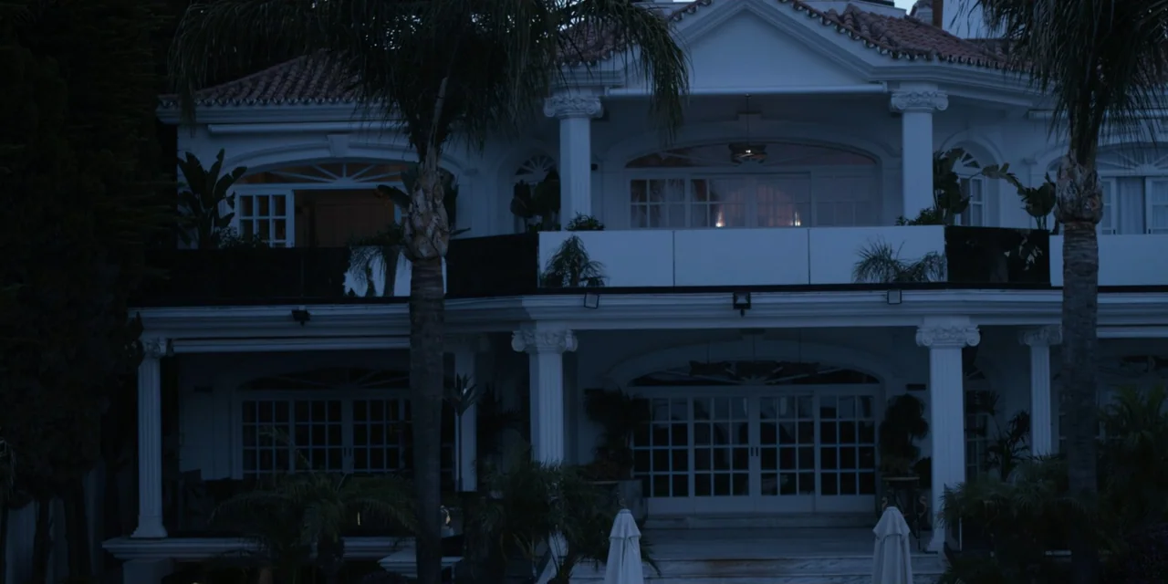 Our Fault (2025) movie still: establishing — A two-story white mansion features a central palm tree and neoclassical architectural pillars. The…