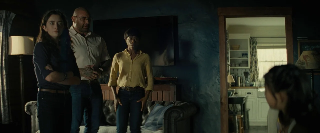 Knock at the Cabin (2023) movie still: medium — A dimly lit room with dark blue walls contains a tufted leather sofa. Three adults stand in the…