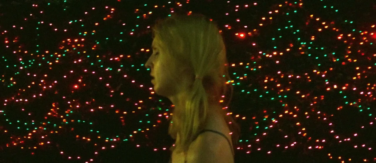 Tangerine (2015) movie still: medium — A dark indoor space is filled with hundreds of tiny, blurred red and green lights. A young woman with blonde…
