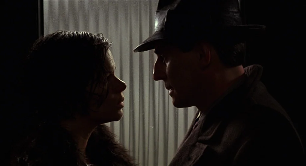 Miller’s Crossing (1990) movie still: medium — A dark, dimly lit room contains two figures facing one another before a window. The woman with dark…