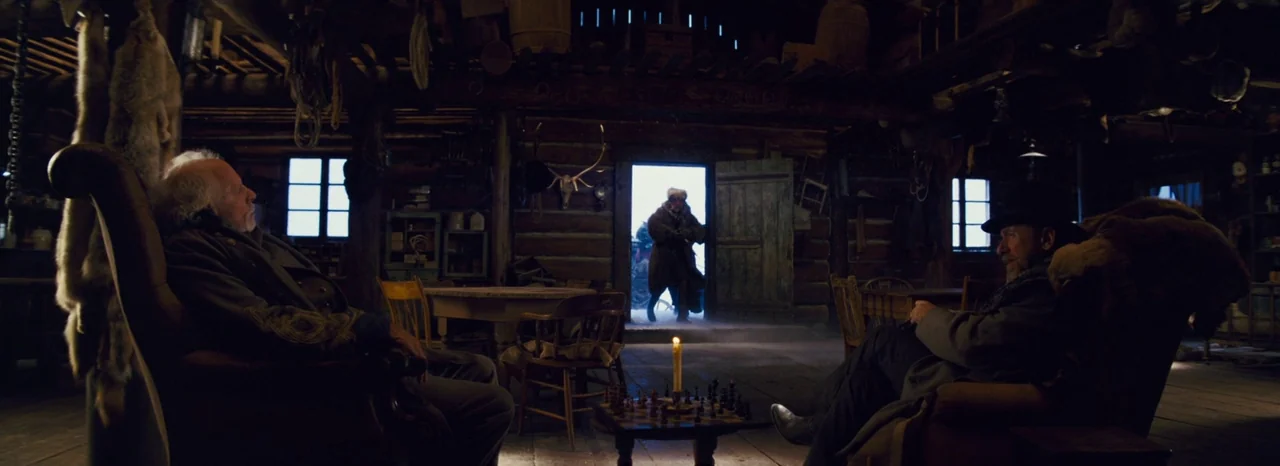 The Hateful Eight (2015) movie still: wide — A dark wooden log cabin filled with scattered animal pelts and rustic furniture. Three men remain…