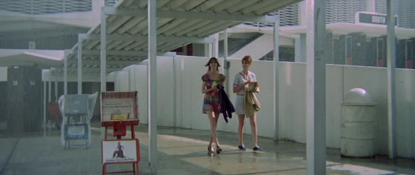 Brewster McCloud (1970) movie still: fullbody — An outdoor concrete walkway with a white roof canopy and modernist architectural details. Two women…
