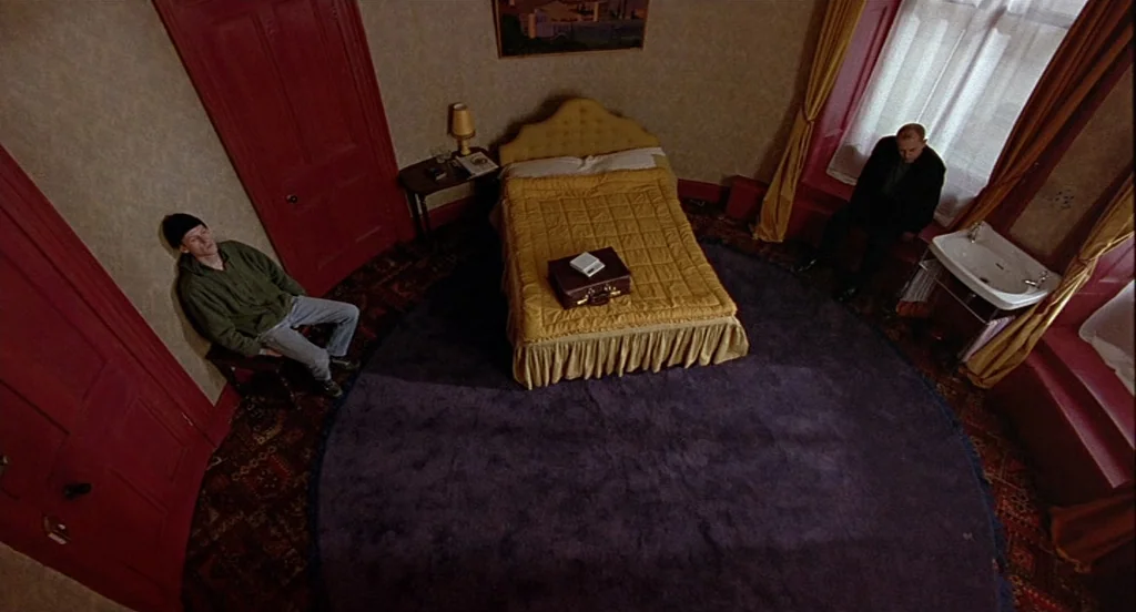 Trainspotting (1996) movie still: wide — A hotel room features red walls and a large round purple carpet in the center. A man sits in a chair near…