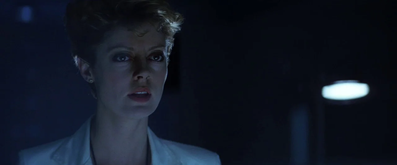 The Hunger (1983) movie still: closeup — A dark, dimly lit professional environment with deep blue shadows. A woman with short wavy hair looks…
