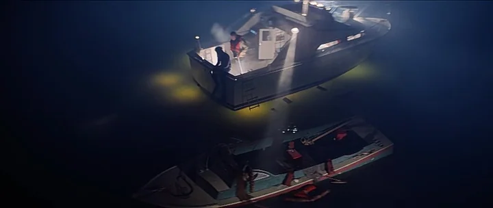 Jaws movie still: wide — Two boats float on the dark, calm surface of the open water at night. The larger boat projects a powerful beam of light…