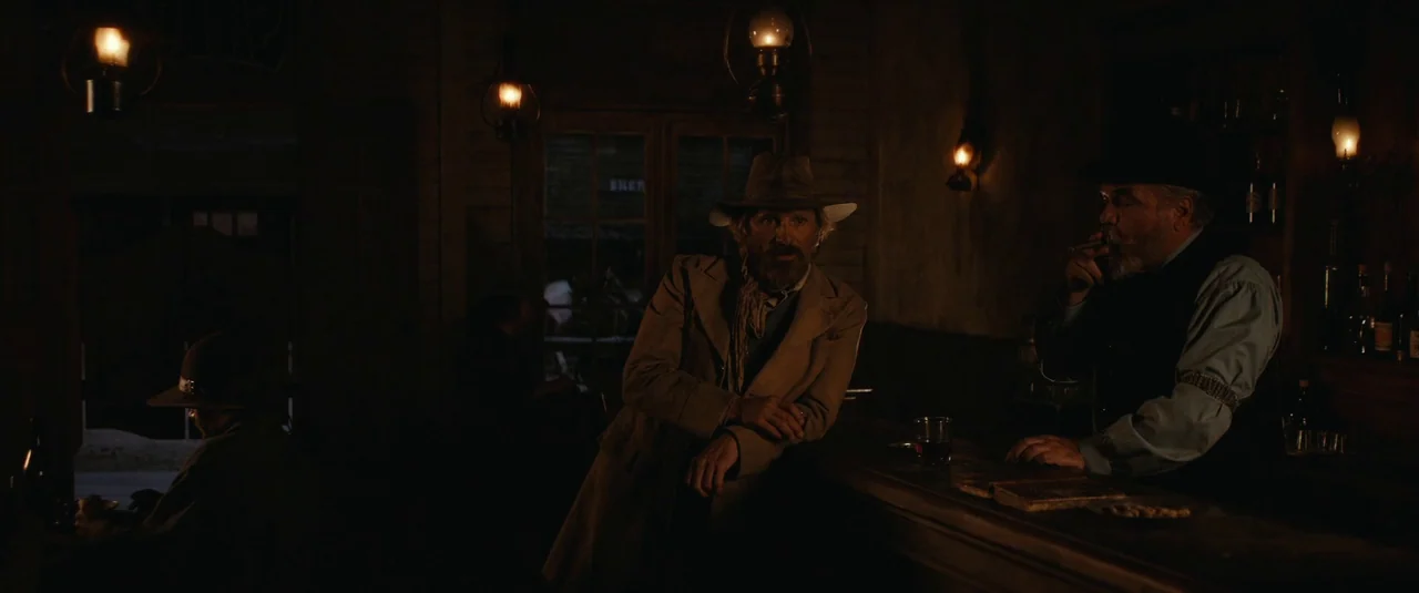 The Dead Don’t Hurt (2023) movie still: medium — A dimly lit old wooden establishment featuring warm oil lamp light. Two men stand at the counter in…